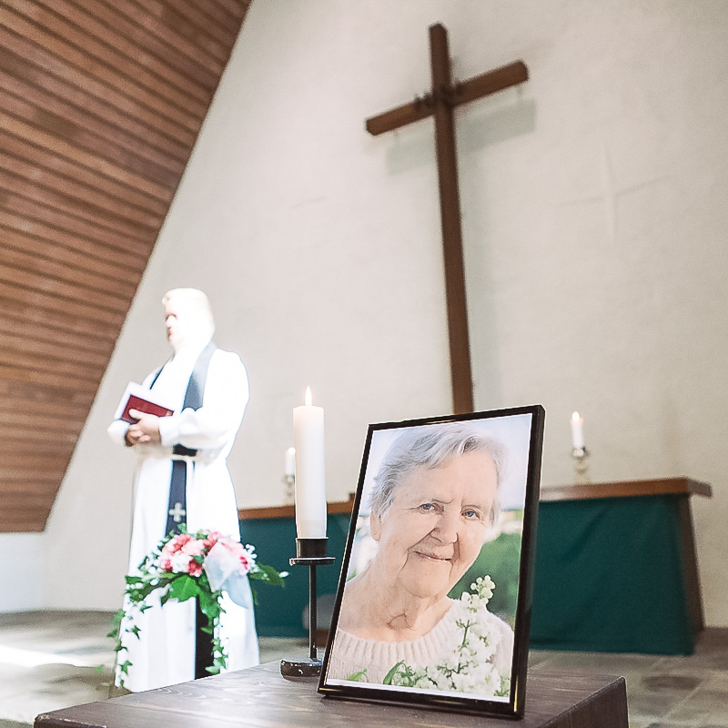 Vainajan valokuva kirkossa ja taustalla pappi.
